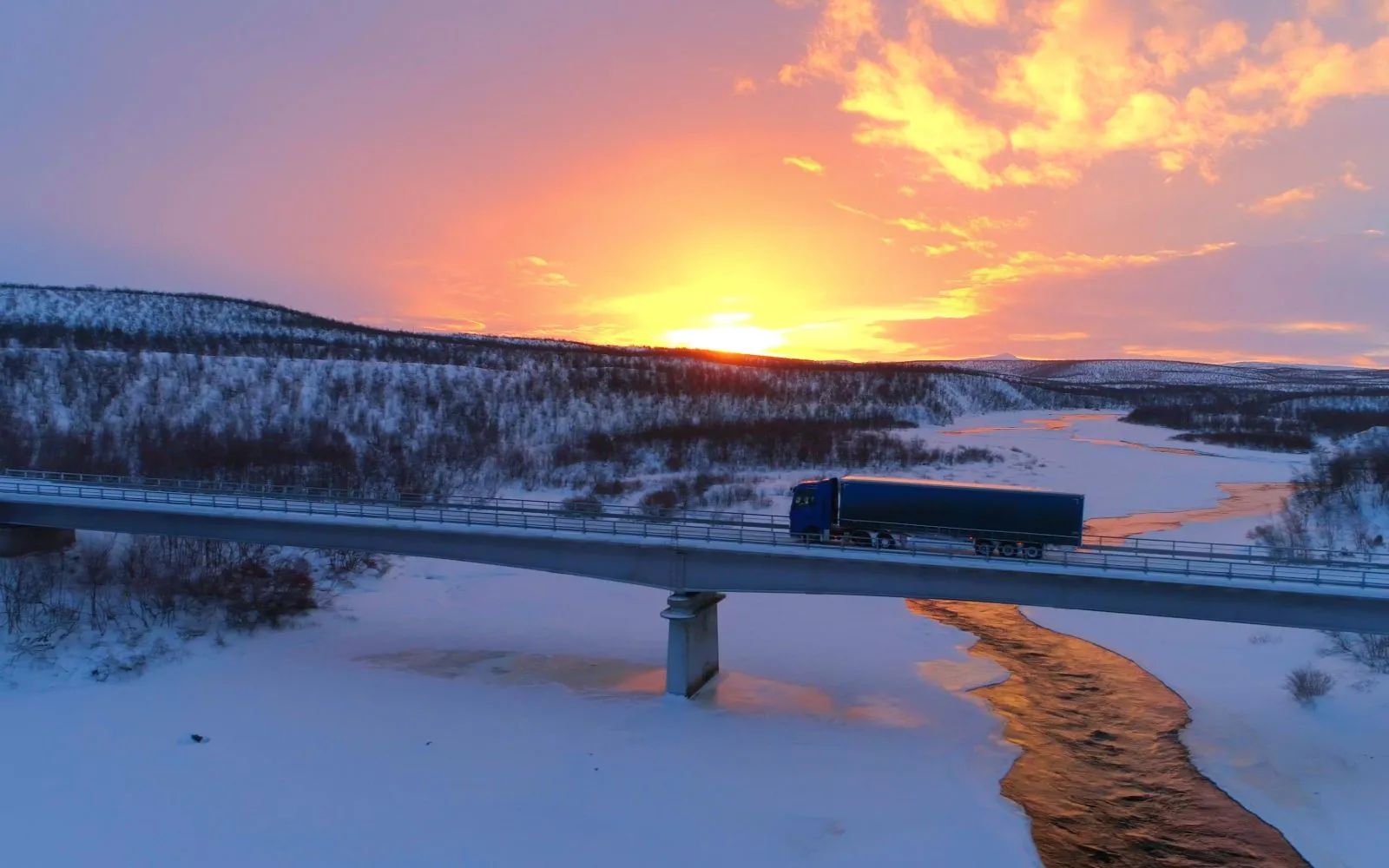 Lastebil på bro over et snødekt landskap i solnedgang